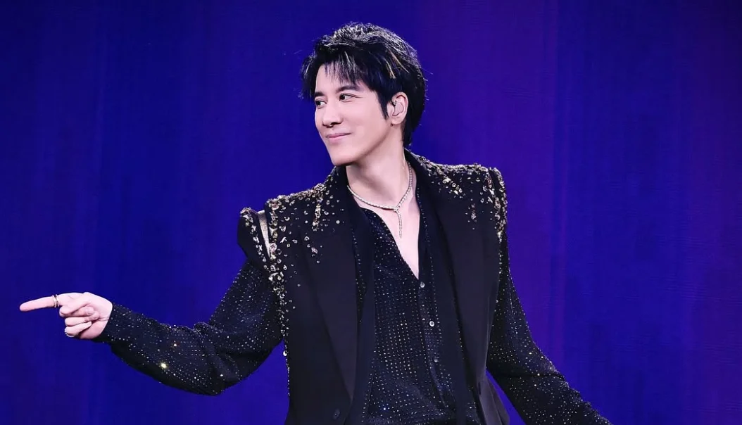 Wang Leehom on stage wearing a black, sparkly outfit with gold embellishments, smiling and pointing to the side against a purple backdrop