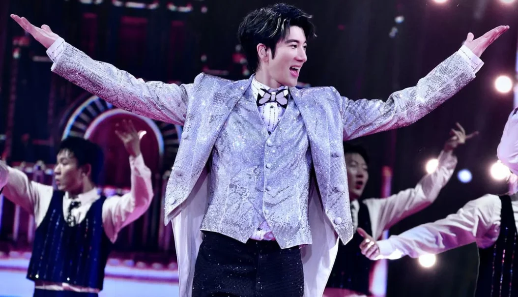 Wang Leehom in a sparkling silver suit spreads his arms wide while smiling on stage, with background dancers in coordinated outfits behind him under bright stage lights