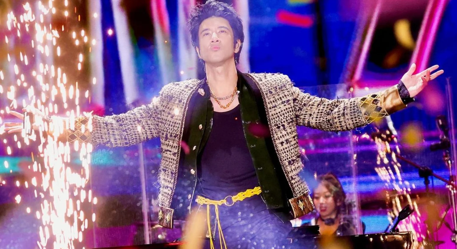 Wang Leehom performing on stage with arms wide open, colorful lights and sparks creating a dramatic concert atmosphere