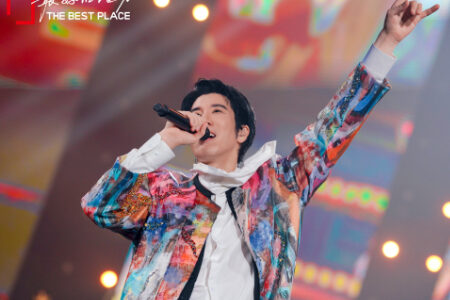 Wang Leehom performing energetically in a vibrant colorful tie-dye jacket with one arm raised on stage