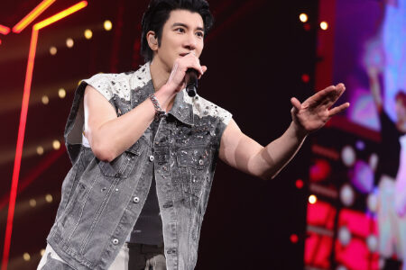 Close-up of Wang Leehom in denim vest holding microphone with warm bokeh stage lights behind him