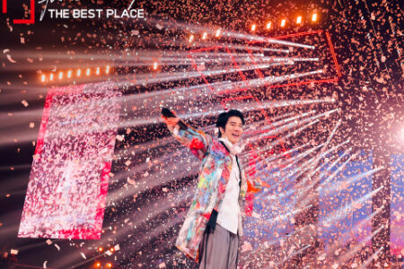Wang Leehom in colorful tie-dye jacket with arm raised triumphantly surrounded by dramatic water spray effects on stage